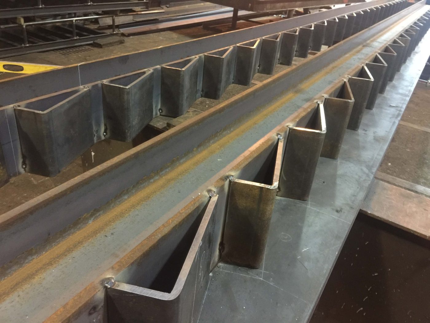Two long steel stringers lined with evenly spaced bent-plate brackets tack welded along their length, receding into a fabrication shop.