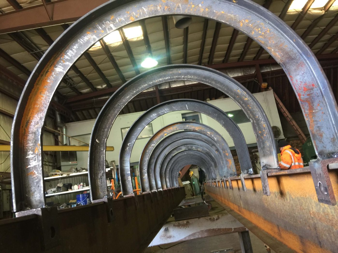 A receding row of curved rolled steel arch ribs standing on a long beam inside a fabrication shop, with a worker in an orange hi-vis jacket at the right.