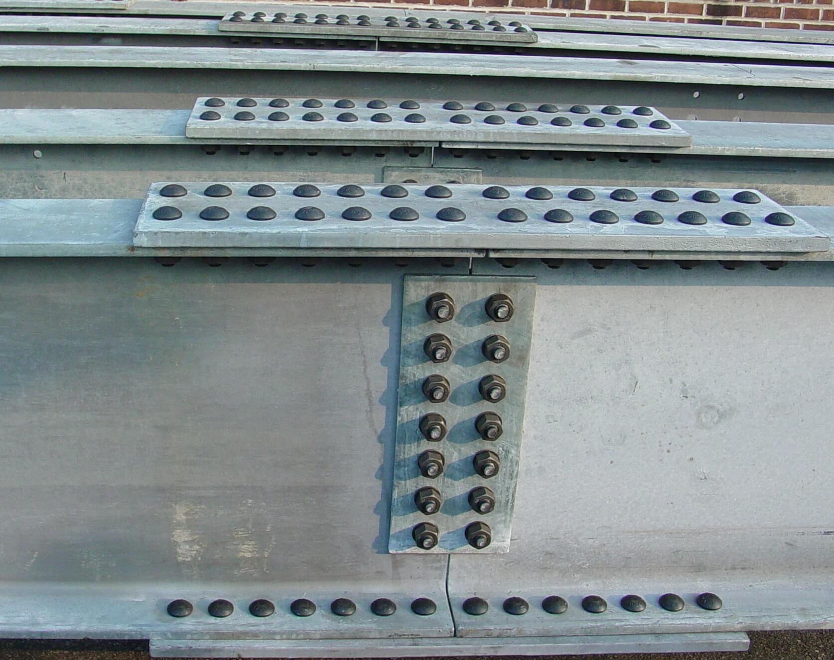 Close view of a galvanized steel girder splice with a bolted web plate and bolted flange cover plates, rows of high-strength bolt heads visible.
