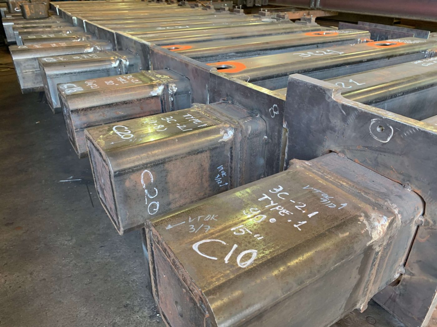 A row of welded steel box columns lying on a shop floor, their capped ends chalk marked with piece numbers and type callouts.