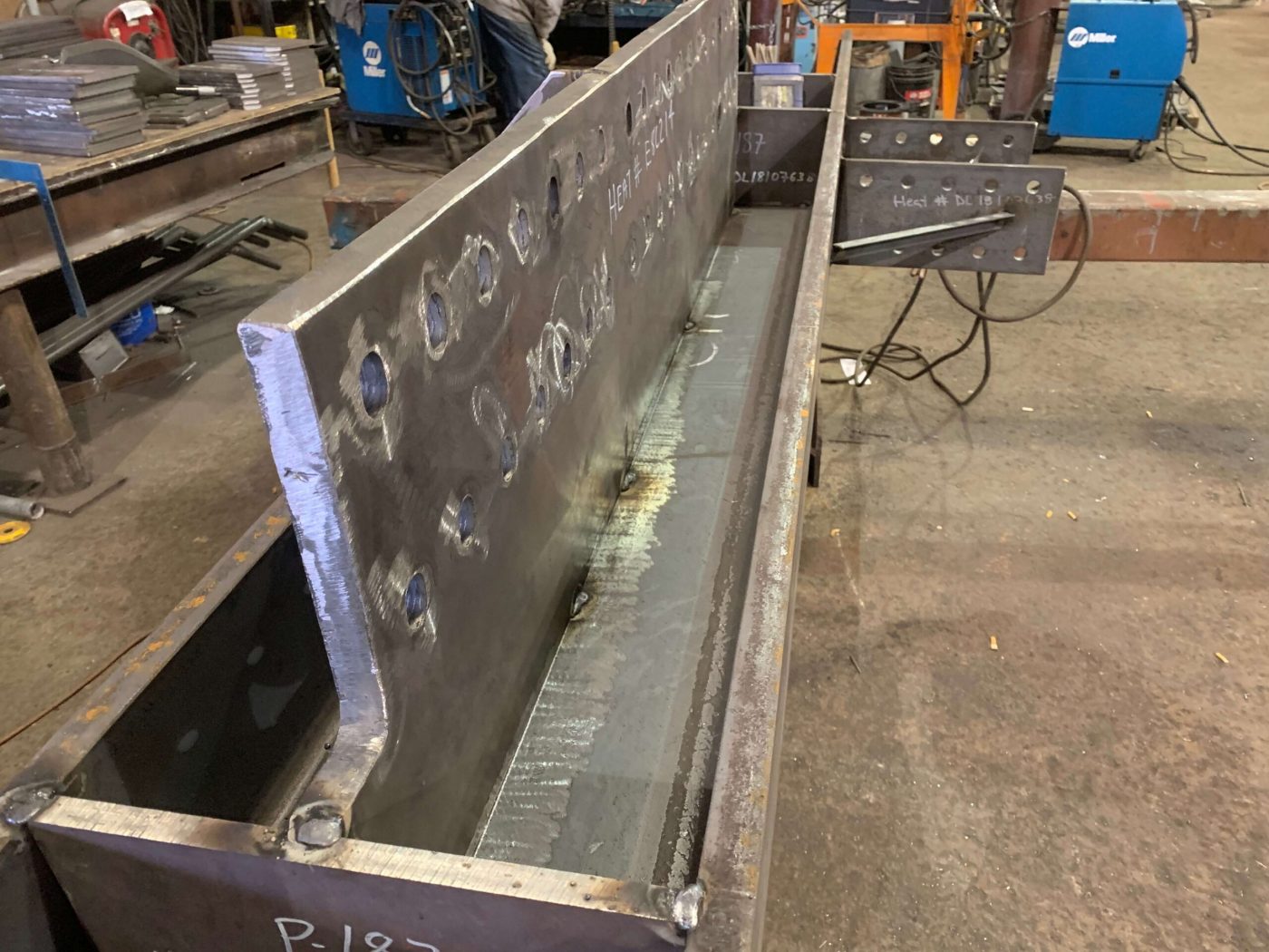 A long welded built-up steel box beam with a beveled edge and chalked heat numbers sits on a fabrication shop floor, welding machines lined up behind it.