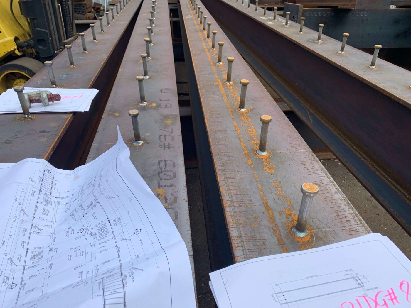 Steel beams laid side by side with rows of shear studs welded along their top flanges, shop drawings and a lifting shackle resting on the steel.