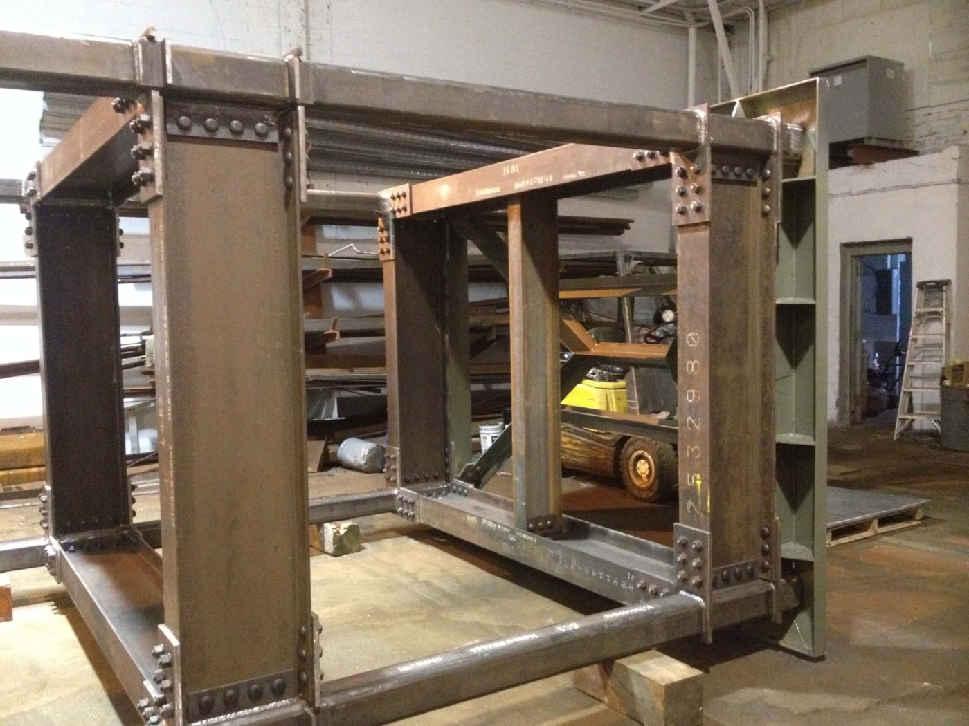 A large rectangular steel frame assembled with rows of heavy domed bolt heads at each corner connection, standing on timber blocking inside a fabrication shop.