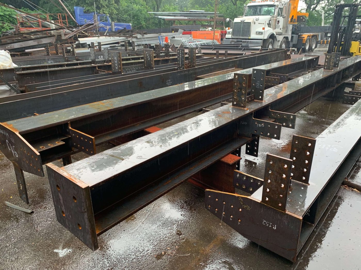 Rows of finished steel beams and columns with bolted connection plates stacked in a rain-wet fabrication yard, a boom truck and forklift in the background.