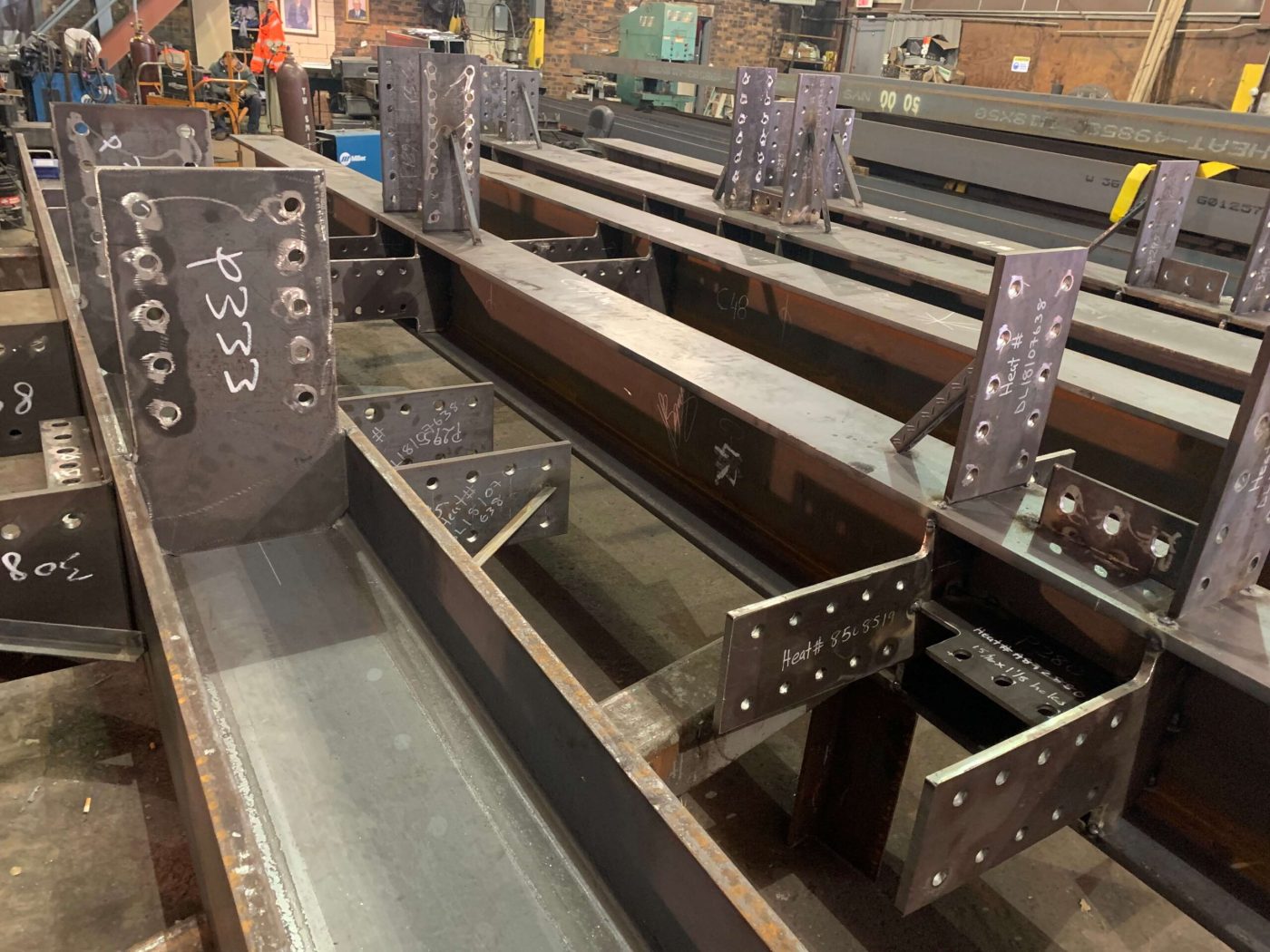 A fabrication shop bay filled with rows of steel framing beams fitted with drilled connection plates, with a work bench and machinery at the far end.