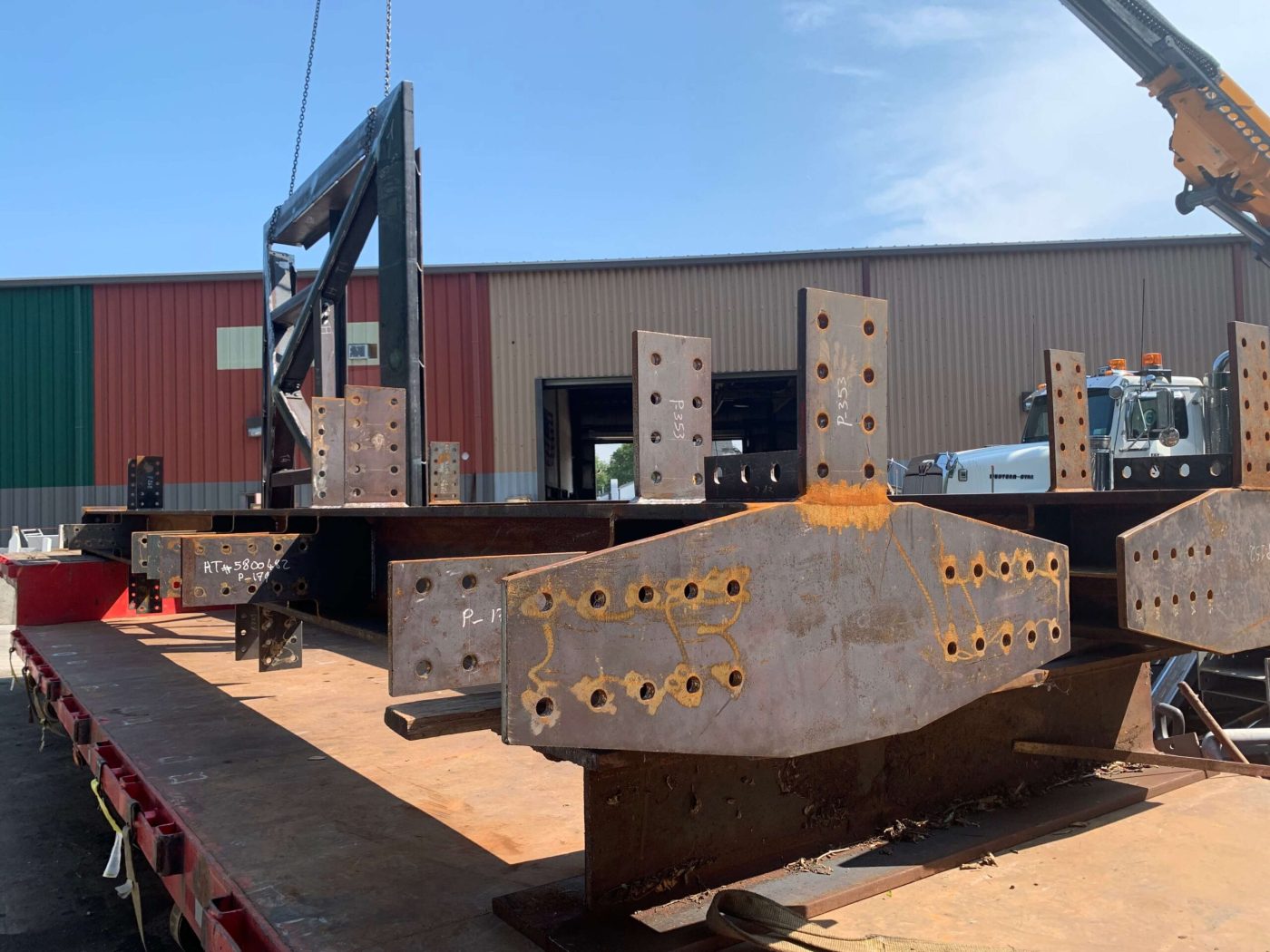 Fabricated steel members with drilled connection plates loaded on a red flatbed trailer outside a fabrication shop, a boom crane rigged above under a clear blue sky.