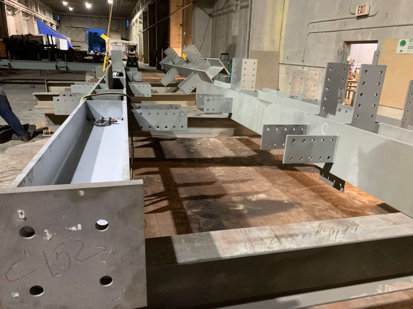 A long row of primed grey steel beams with drilled connection plates stretching down a fabrication shop floor.