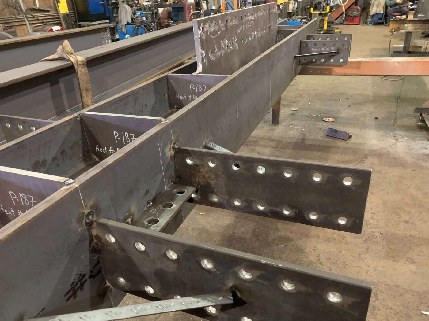 Raw steel girders with welded stiffeners and bolted splice plates on a shop floor, piece marks and heat numbers written in chalk.