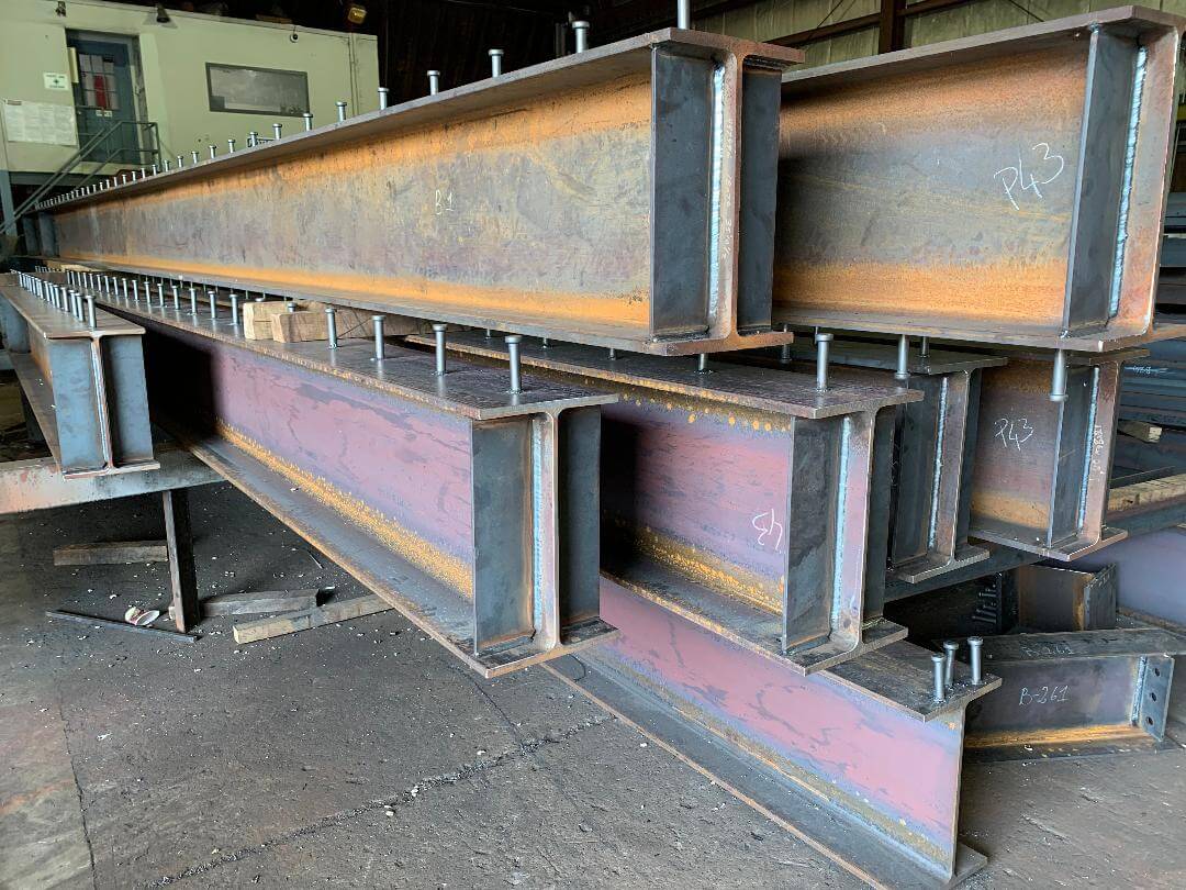 Long steel girders resting on blocking in a fabrication shop with rows of welded shear studs running along the top flanges.