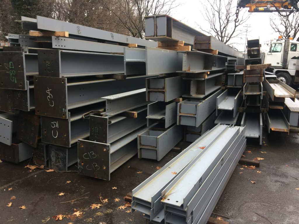 A yard stack of primed steel beams, columns and box sections on timber blocking with a boom truck parked alongside.
