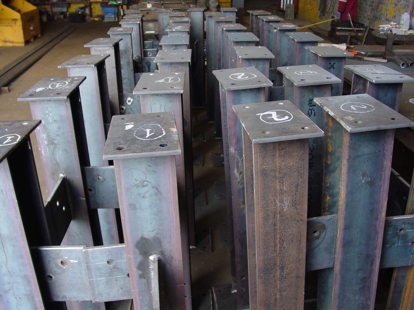 Rows of short steel columns standing upright in a shop, each with a welded cap plate chalk-marked with a piece number.