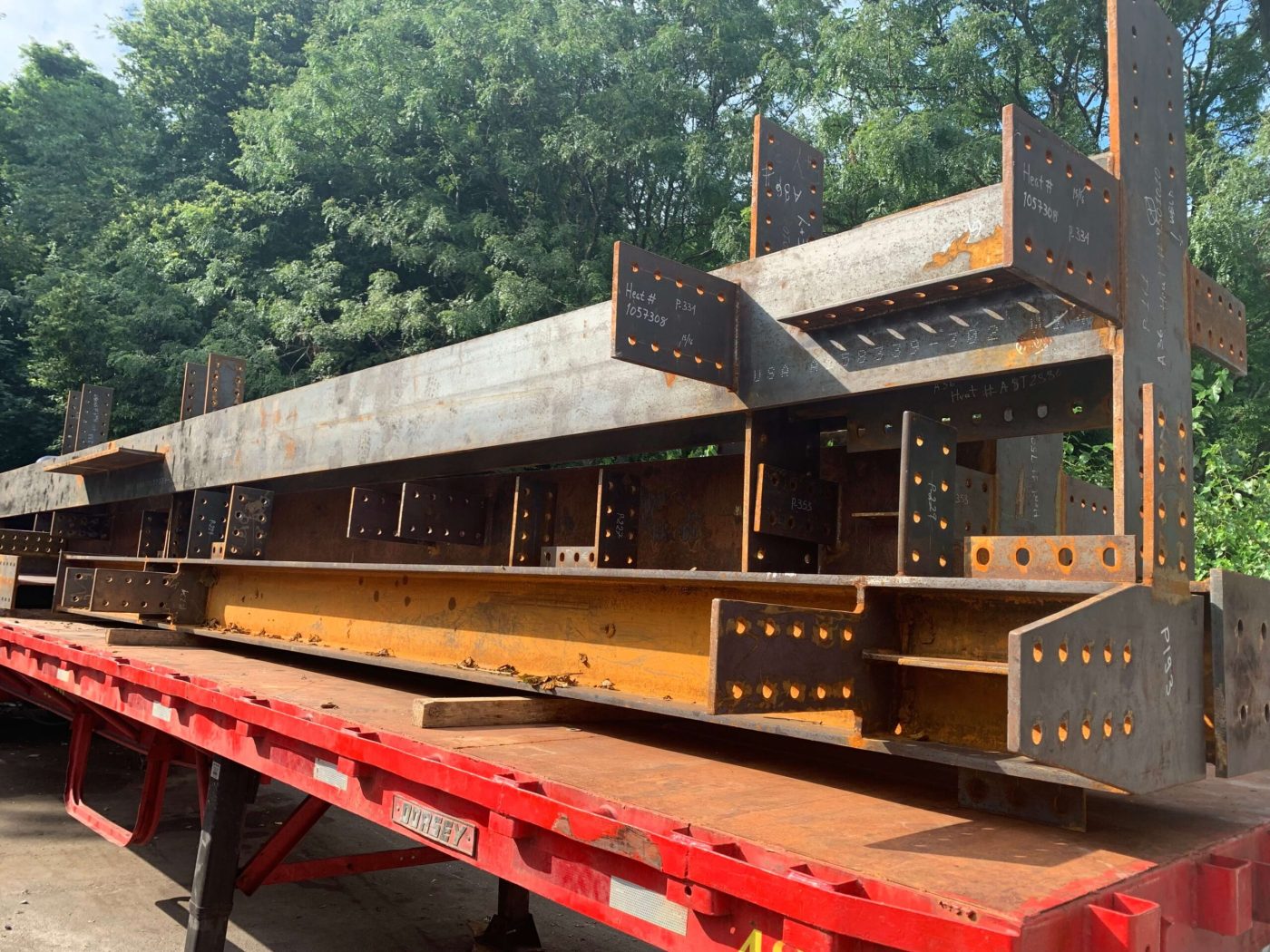 A red flatbed trailer stacked with fabricated steel beams and bolted connection plates marked with white heat numbers, parked in a yard beside green trees.