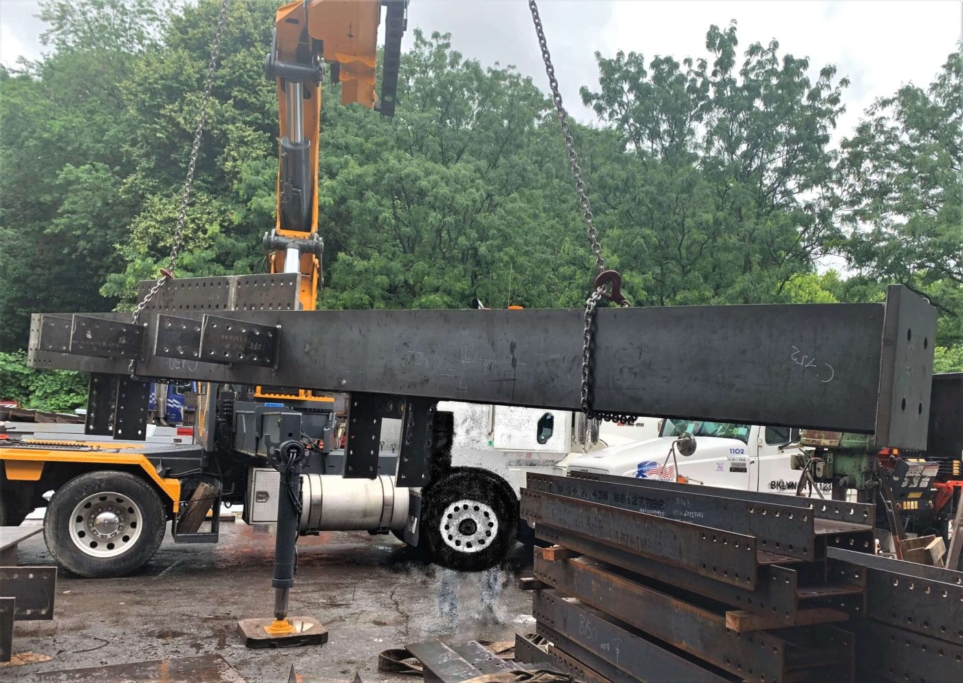 A yellow knuckle-boom truck lifting a long fabricated steel girder on chain slings in a fabrication yard, with stacked steel and trucks behind it.