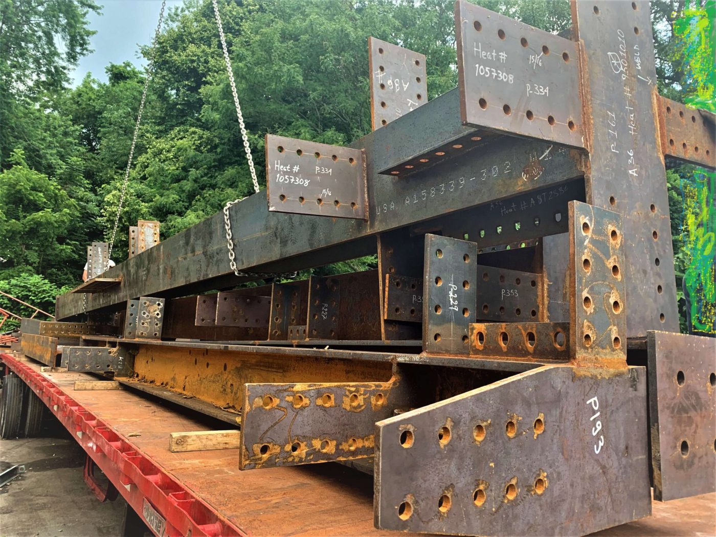 Fabricated steel beams and connection plates stacked on a red flatbed trailer, with heat numbers and piece marks written in white on the plates.