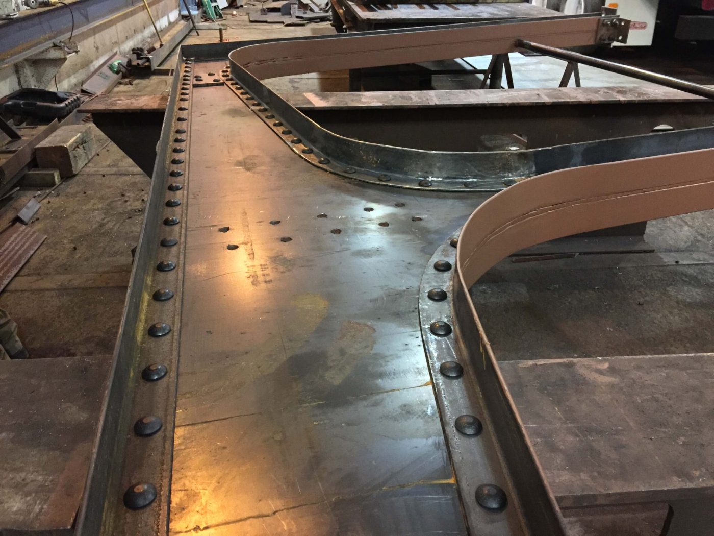 Curved built-up steel girder sections laid out on a fabrication shop floor, their flange plates fastened with long rows of heavy hex bolts.