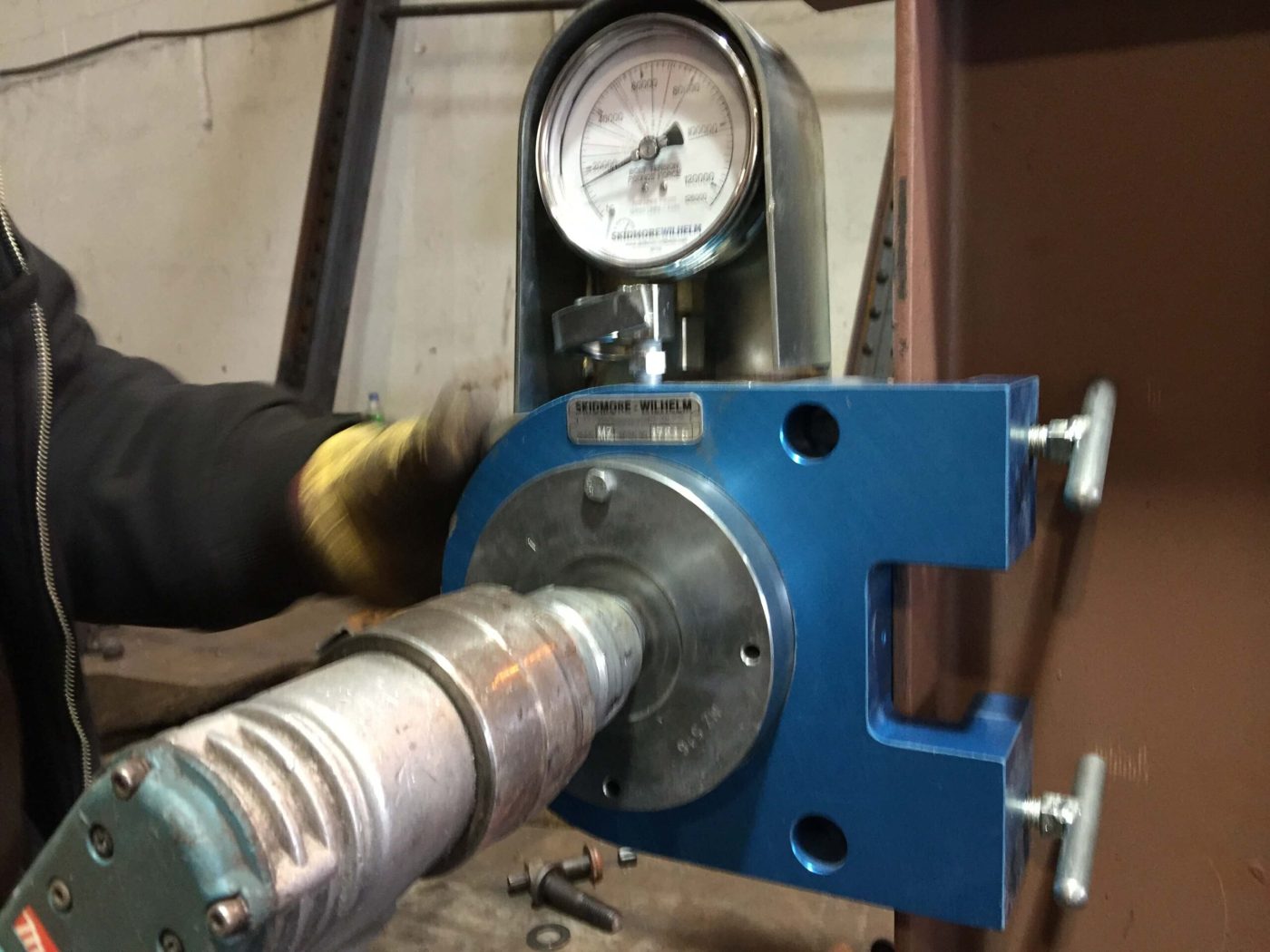 A blue Skidmore-Wilhelm bolt tension calibrator clamped to a steel flange with an impact wrench driving the test bolt and the pressure dial above it.
