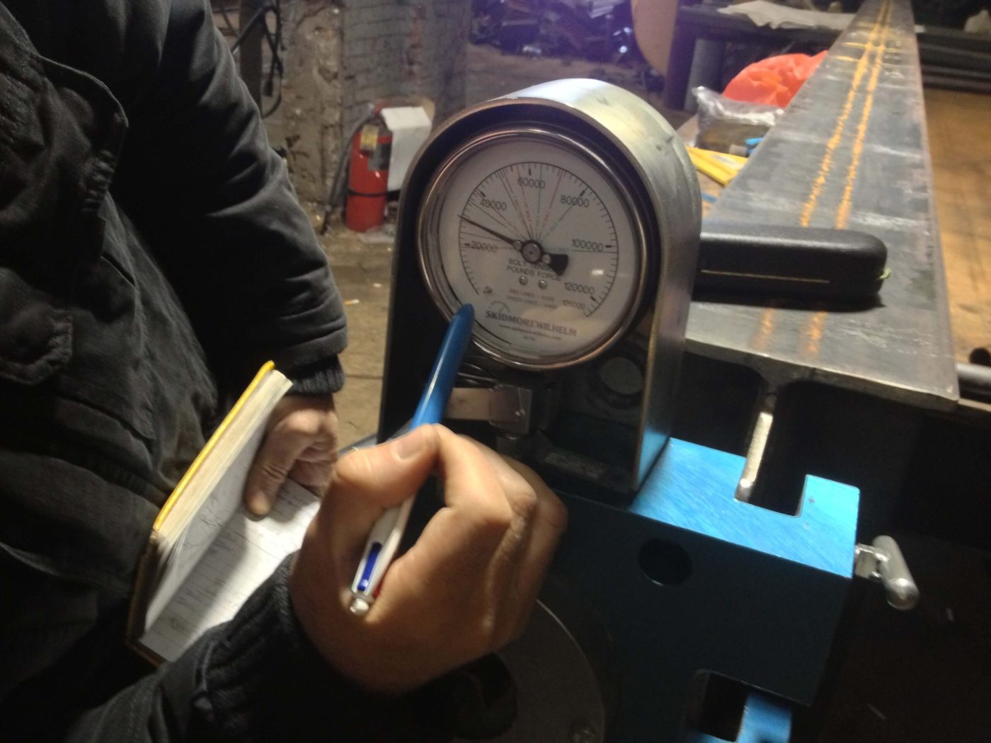 A Skidmore-Wilhelm bolt tension gauge sits on a shop bench while a hand writes the reading into a small field notebook.