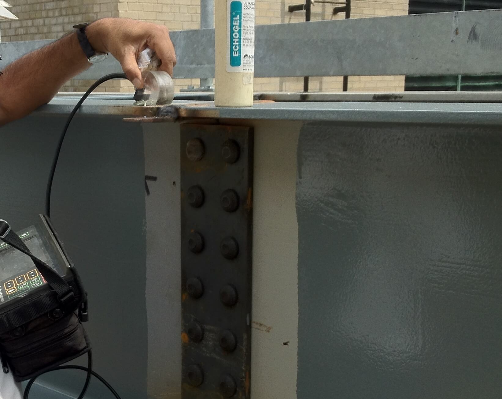 A hand holds an ultrasonic probe against the top of a painted steel girder beside a bolted splice plate, with couplant gel and a handheld flaw detector nearby.
