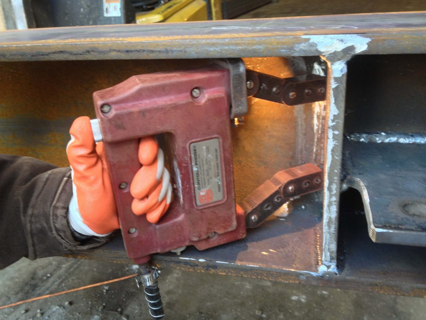 An orange-gloved hand presses a red magnetic particle yoke with jointed legs against a ground weld between a stiffener plate and a beam web.