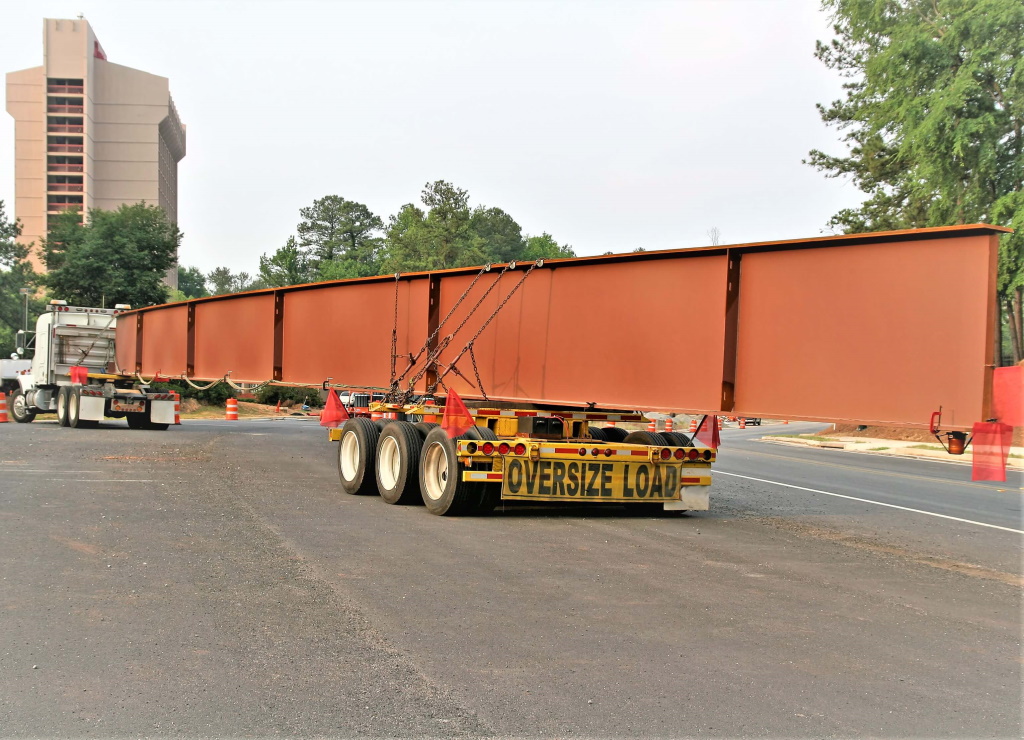 A long red-oxide primed steel plate girder is chained to an extendable drop trailer marked OVERSIZE LOAD, stretching the width of the frame on a paved road with trees and a tower behind.
