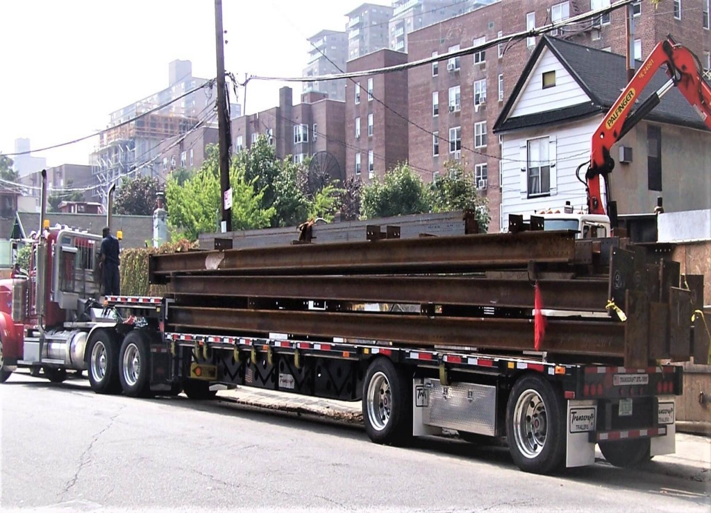 A flatbed trailer stacked with mill-scale steel beams is parked along a city street, a red tractor at its head and a knuckle-boom crane truck alongside, with brick apartment blocks and overhead wires behind.