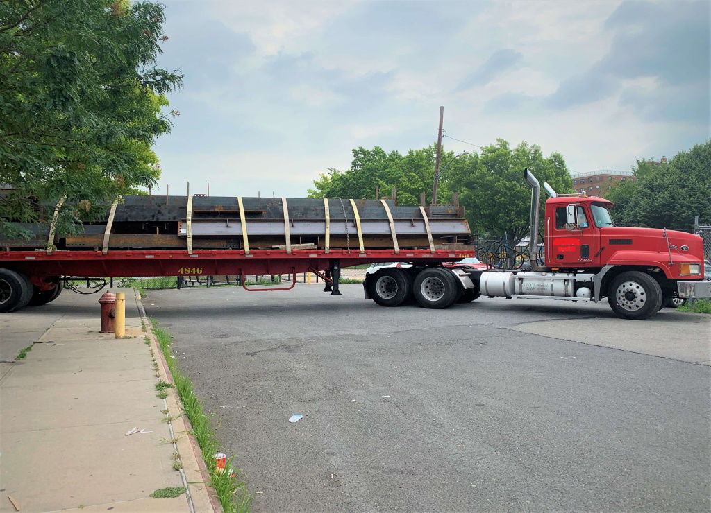 A red Mack tractor pulls a red flatbed trailer carrying strapped steel beams across the frame, parked on an open asphalt lot with trees and a sidewalk in front.