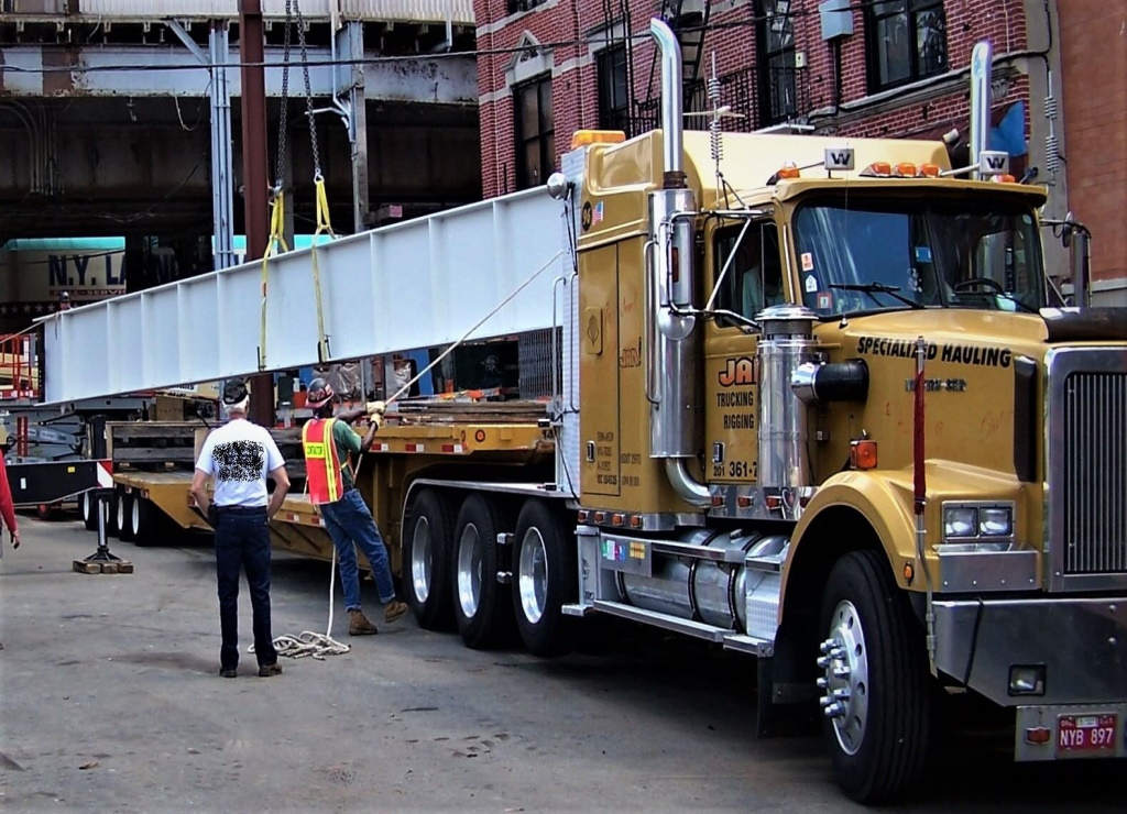 Two workers, one in a hi-vis vest holding a tag line, guide a long white-painted steel plate girder slung on straps as it is loaded onto a yellow heavy-haul tractor and lowboy trailer between city buildings.