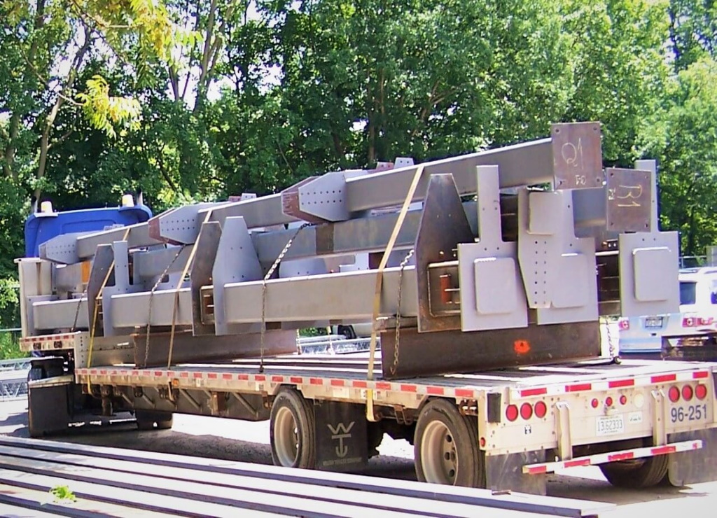 Fabricated steel girders with bolt-holed gusset and connection plates, chalk piece marks visible, sit chained and strapped on a flatbed trailer with green trees behind.