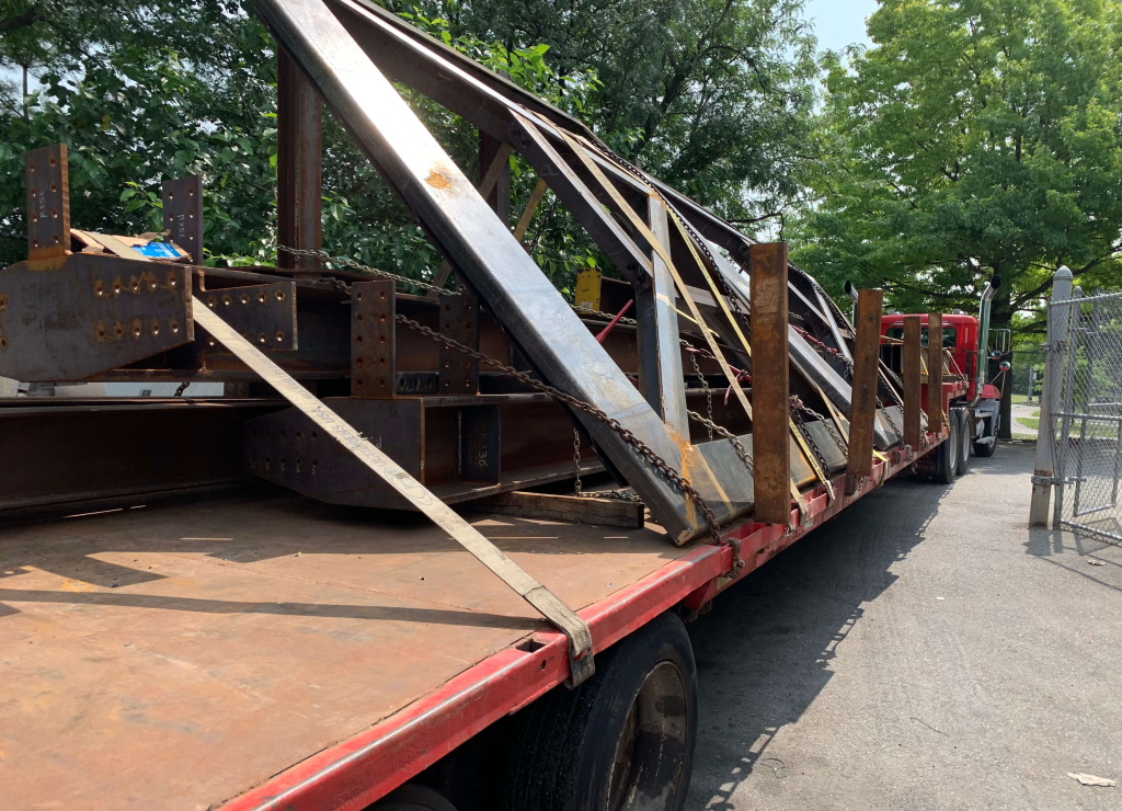 Long fabricated steel trusses and beams with bolted splice plates lean chained and strapped across a red flatbed trailer, the empty deck running toward the camera and trees overhead.