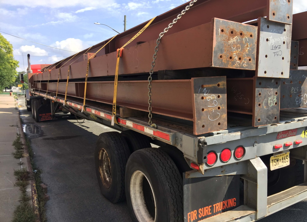 Long red-oxide primed steel beams with chalk-marked bolted end plates run the length of a flatbed trailer, held by yellow straps and a chain, parked on a street under a blue sky.