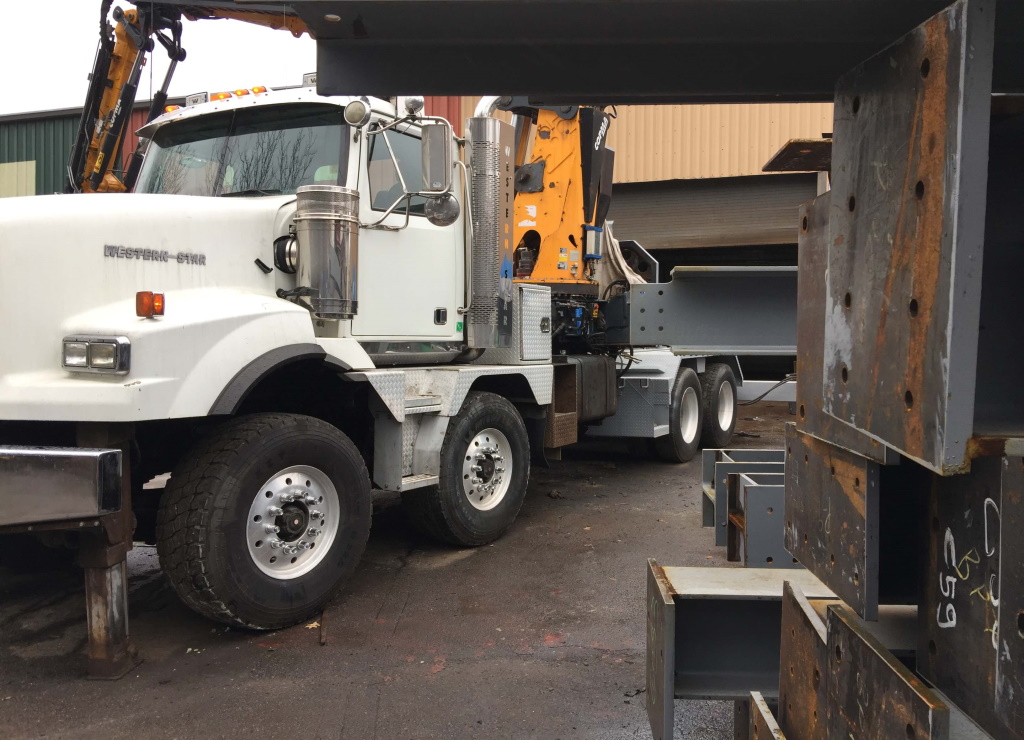 A white heavy tractor fitted with a knuckle-boom crane stands in a fabrication yard under an overcast sky, with chalk piece-marked steel members stacked in the right foreground.