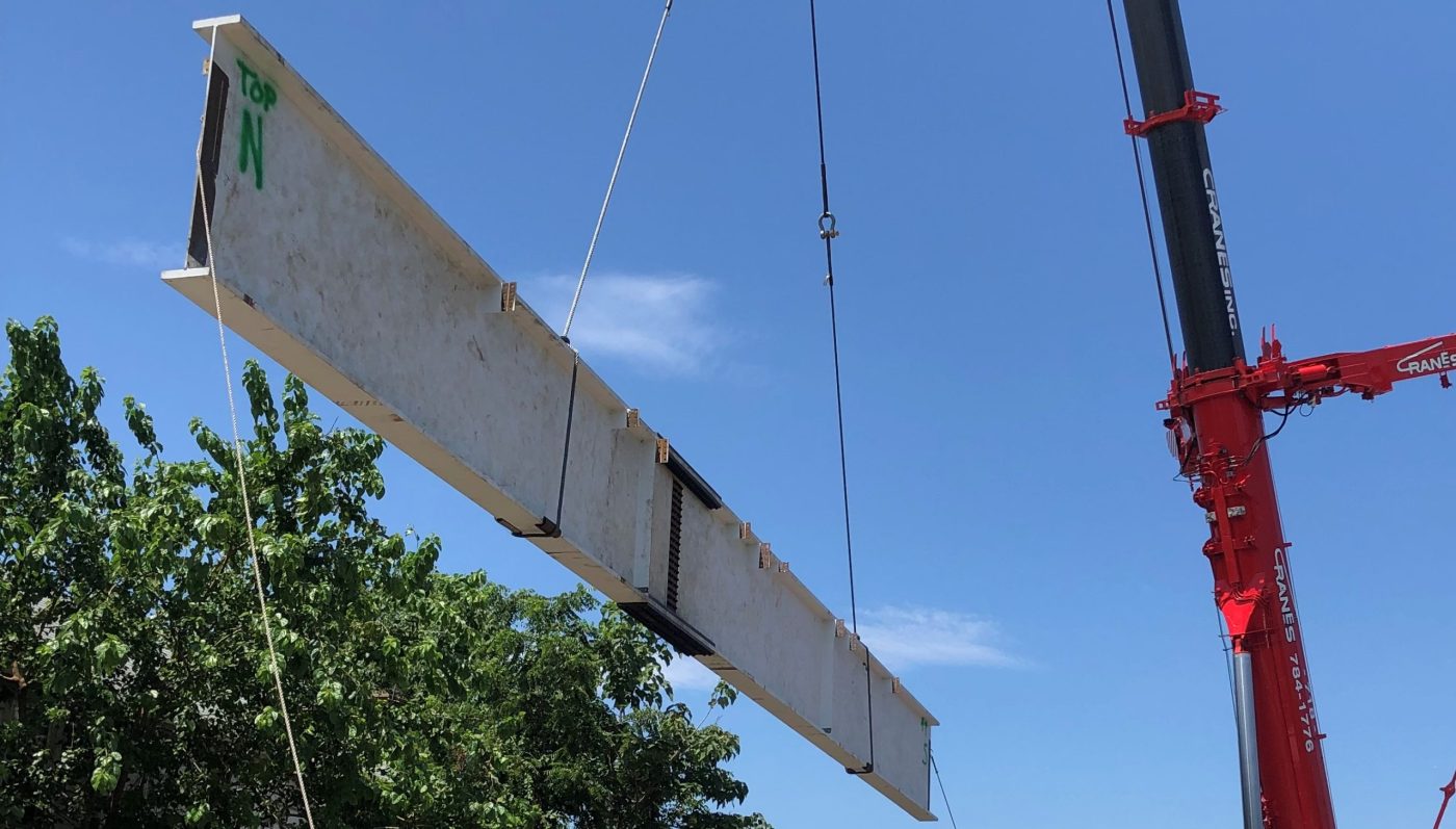 A long white-primed steel plate girder marked TOP in green paint hangs diagonally from a red mobile crane against a blue sky.