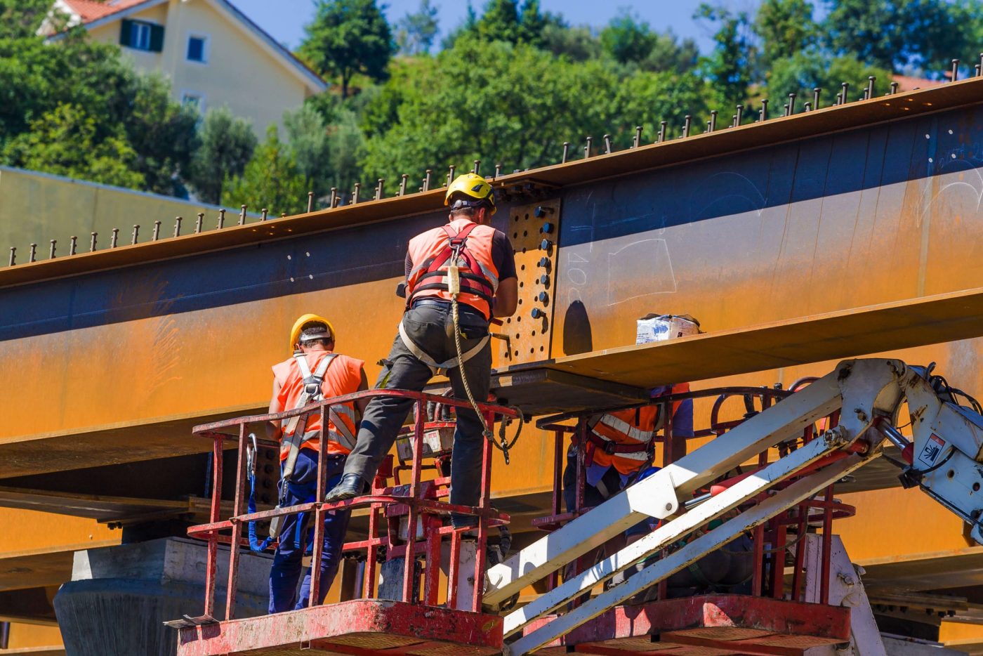 Two ironworkers in orange vests and hard hats standing on a lift platform to bolt the splice plate of a large bridge girder lined with shear studs.