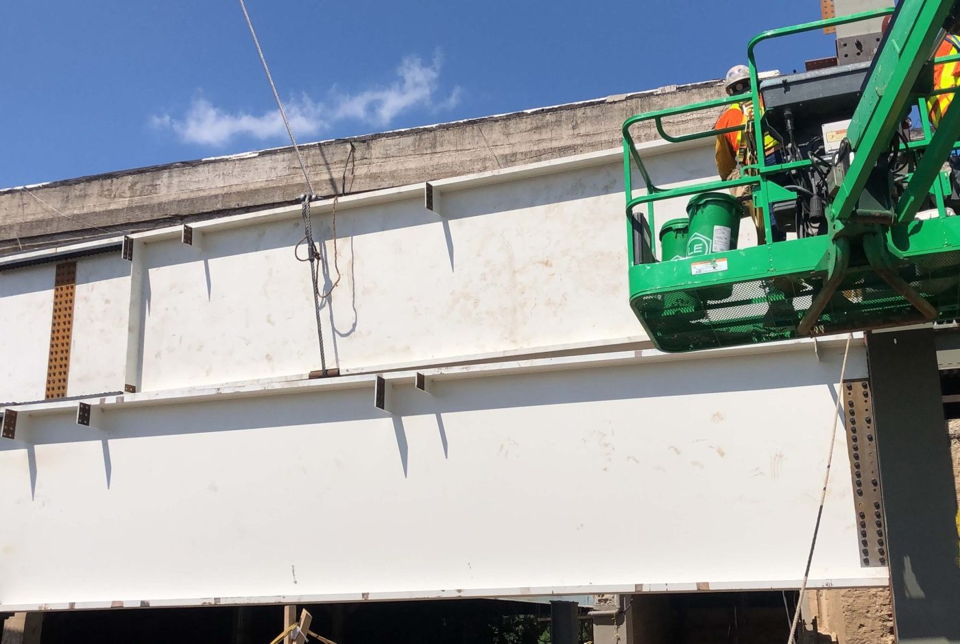 White-painted steel bridge girders with welded stiffeners set beneath a concrete deck, with a green boom lift basket at the right.