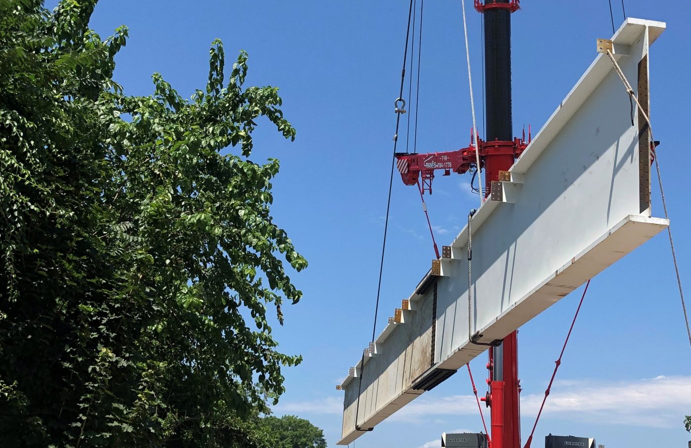A white-primed steel plate girder with rows of connection clips hangs level from wire ropes beside a red mobile crane boom, trees to the left.