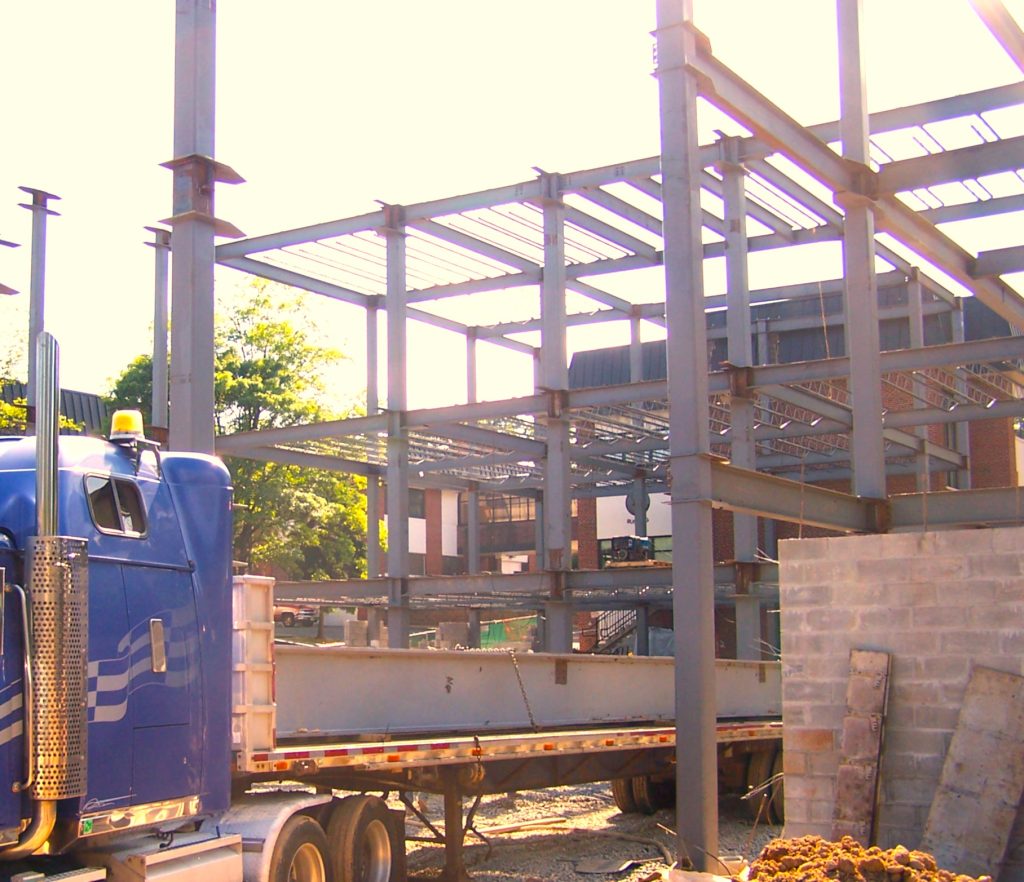 A partly built multi-story steel frame of columns, beams and open-web joists stands against a bright sky, with a blue tractor-trailer loaded with a steel beam parked at the left of the site.