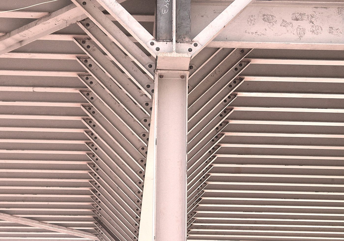 Looking up at a roof structure where two rows of open-web steel bar joists run at an angle into a girder and column connection, with bolt heads visible at every seat.