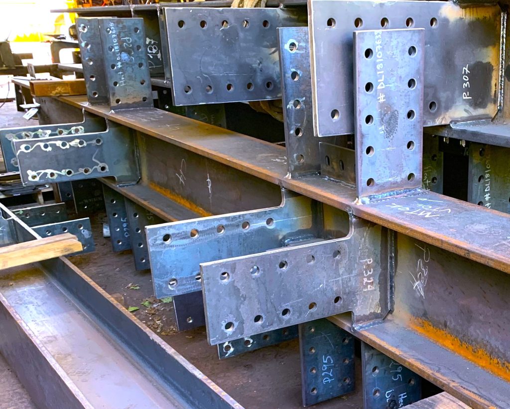 Close view of fabricated steel beams fitted with welded end plates and shear tabs full of bolt holes, heat numbers written on them in chalk.